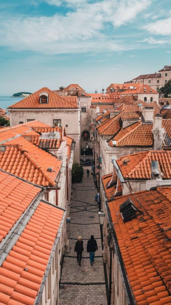  A picturesque view of Dubrovnik's historic streets, lined with orange terracotta rooftops, winding down towards the distant Adriatic Sea under a bright sky. The scene epitomizes the Dubrovnik Croatia aesthetic with its timeless charm.