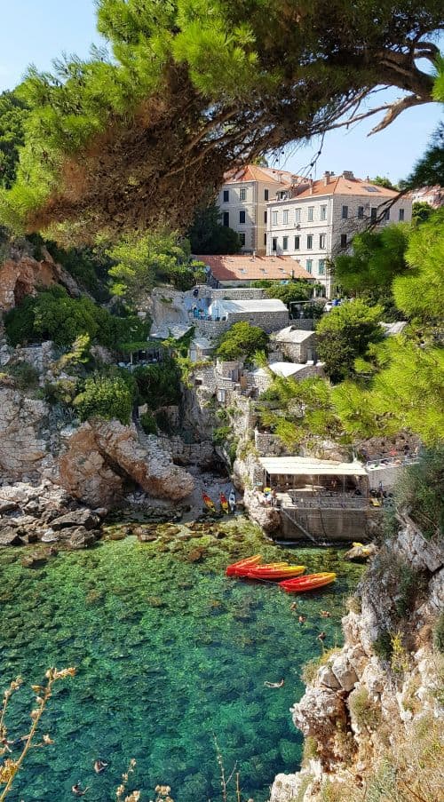  A few red and yellow kayaks float near the shore, with a charming Mediterranean-style building perched on the hillside above. The view is framed by the branches of a pine tree, enhancing the idyllic atmosphere.







