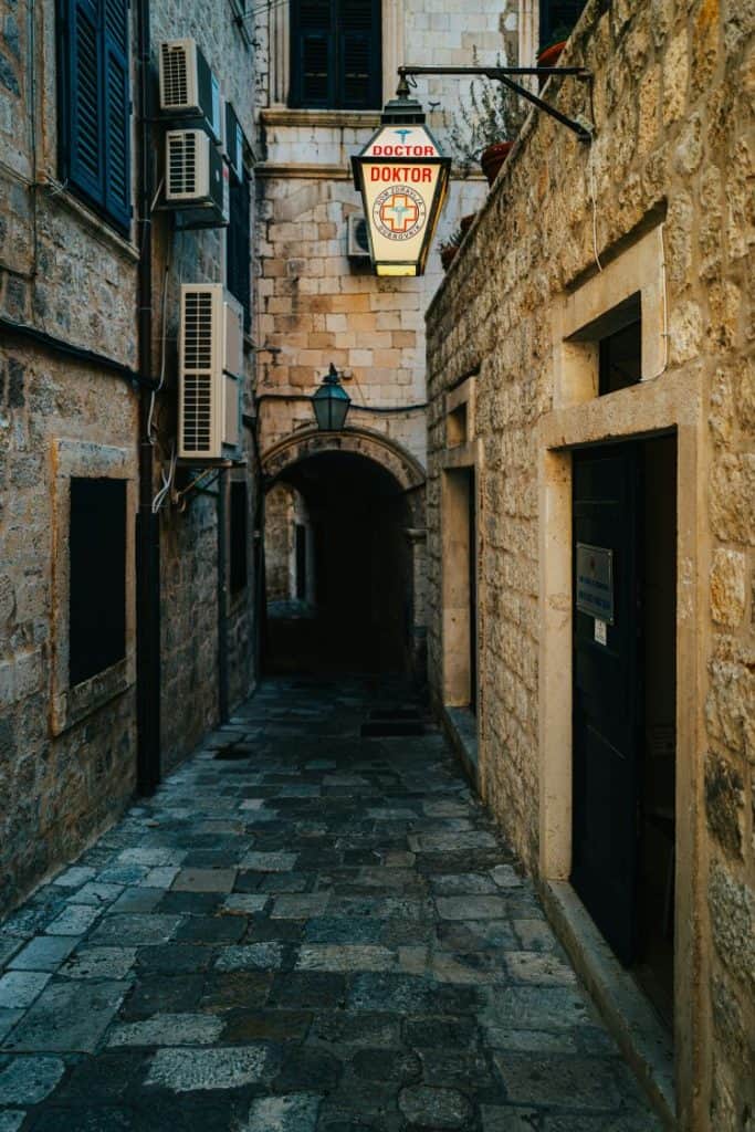  A narrow, cobblestone alleyway in Dubrovnik’s Old Town, bordered by rustic stone walls and a vintage doctor's sign hanging above. The image highlights the historical charm of the Dubrovnik Croatia aesthetic.