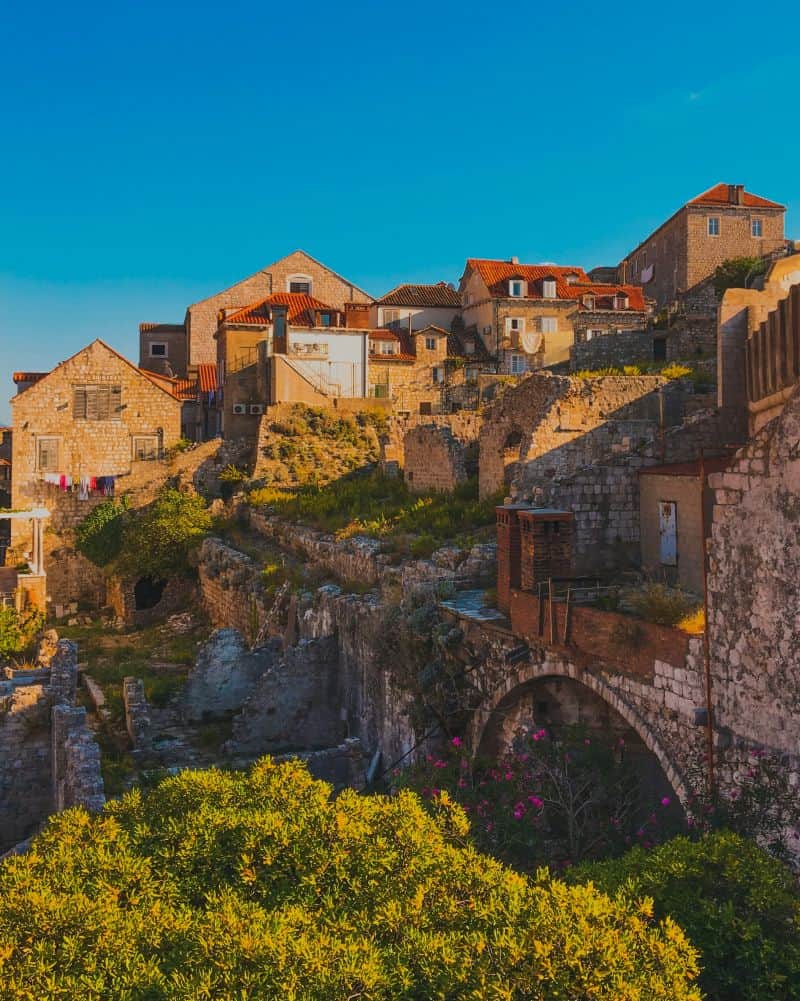A glowing golden-hour view of stone ruins and rustic homes with terracotta rooftops set against a clear blue sky. This warm, idyllic scene embodies the Dubrovnik Croatia aesthetic.