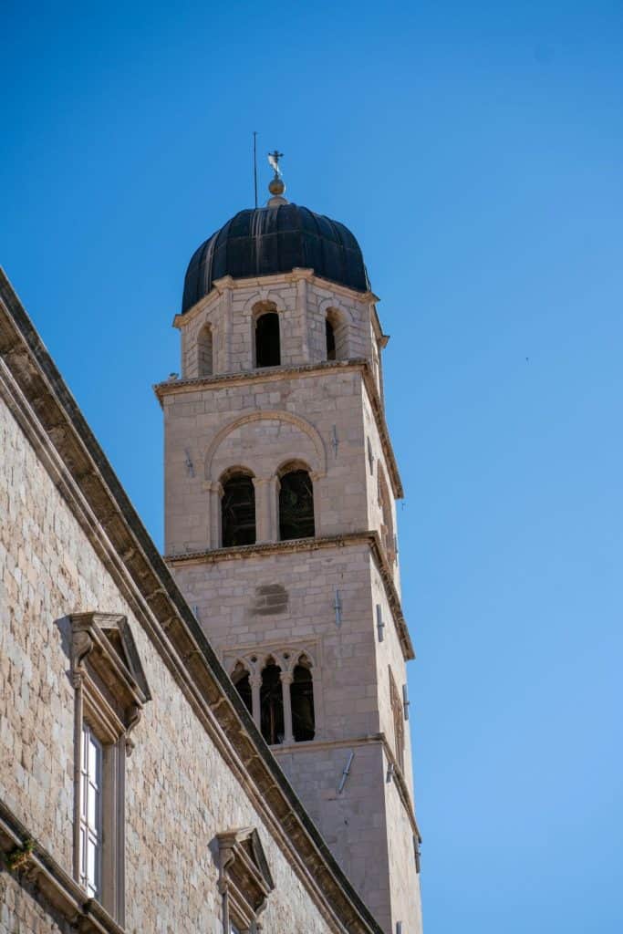  A close-up of a historic bell tower rising against a bright blue sky, showcasing elegant stonework and intricate design. The architectural detail contributes to the Dubrovnik Croatia aesthetic.