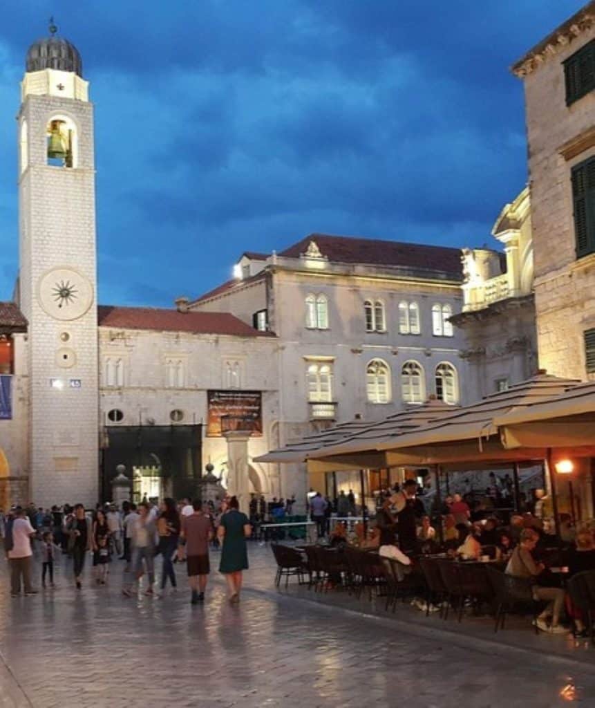 An evening scene in Dubrovnik, Croatia, featuring the illuminated Bell Tower and surrounding historic buildings under a deep blue sky. The lively street is bustling with people walking and dining at outdoor cafes, highlighting the city's vibrant and timeless aesthetic.