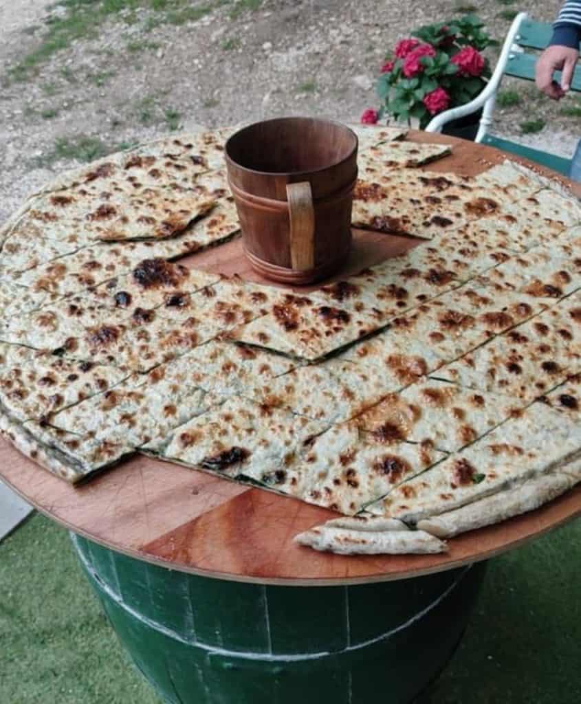 A large, round flatbread served on a rustic wooden table, paired with a traditional wooden bucket in an outdoor setting. The simplicity and tradition of the meal embody the Dubrovnik Croatia aesthetic's culinary heritage.