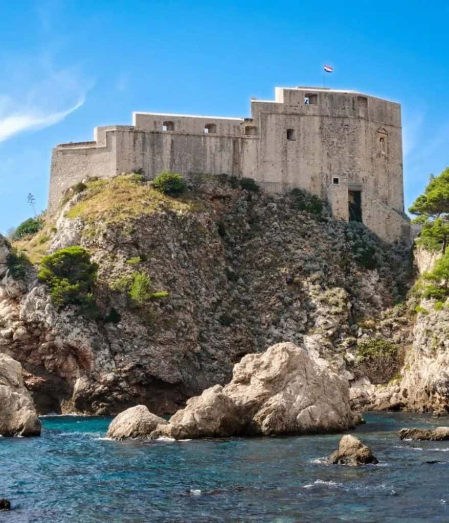 A scenic view of Fort Lovrijenac in Dubrovnik, Croatia, perched on a rocky cliff above the sparkling Adriatic Sea. The fortress, with its stone walls and a Croatian flag atop, is surrounded by rugged terrain and lush greenery, showcasing the aesthetic charm of Dubrovnik's coastal landscape