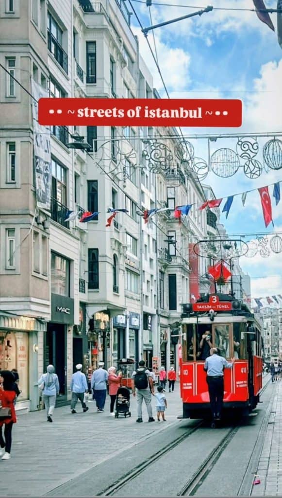 A bustling street in Istanbul featuring the historic red Taksim tram in the center, surrounded by pedestrians, shops, and Turkish flags strung across the street, captured as part of an Istanbul Instagram story.