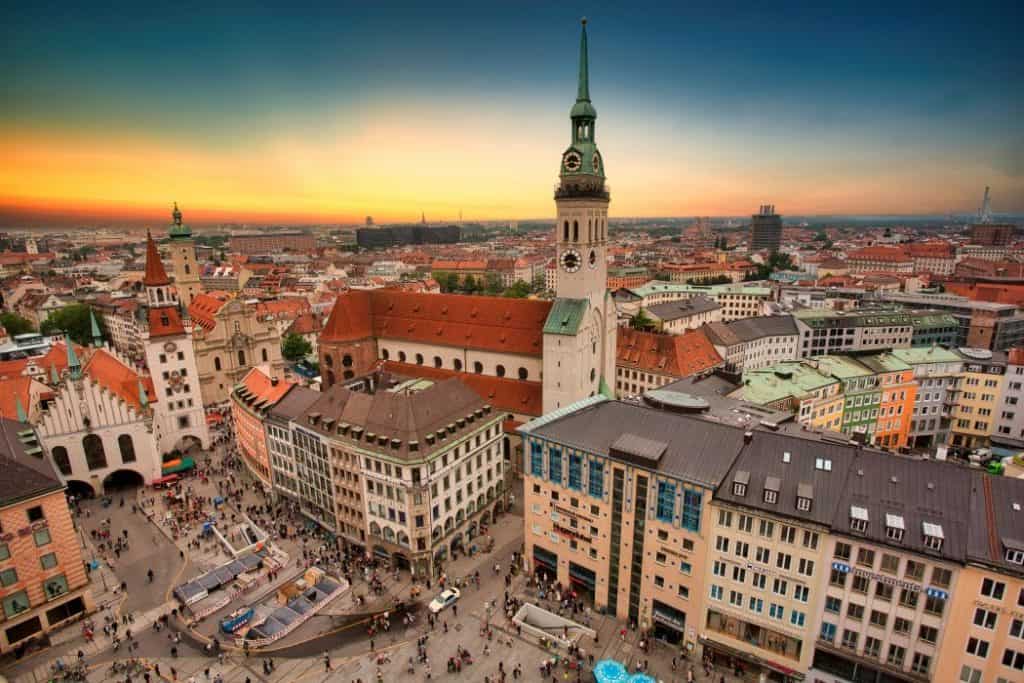  A breathtaking aerial view of Munich at sunset, highlighting St. Peter's Church (Alter Peter) with its tall clock tower—one of the top landmarks among things to do in Munich. The vibrant Marienplatz below is bustling with people, surrounded by historic buildings with colorful facades and red-tiled roofs. The dramatic sky, fading from warm orange to deep blue, enhances the charm of this lively cityscape.