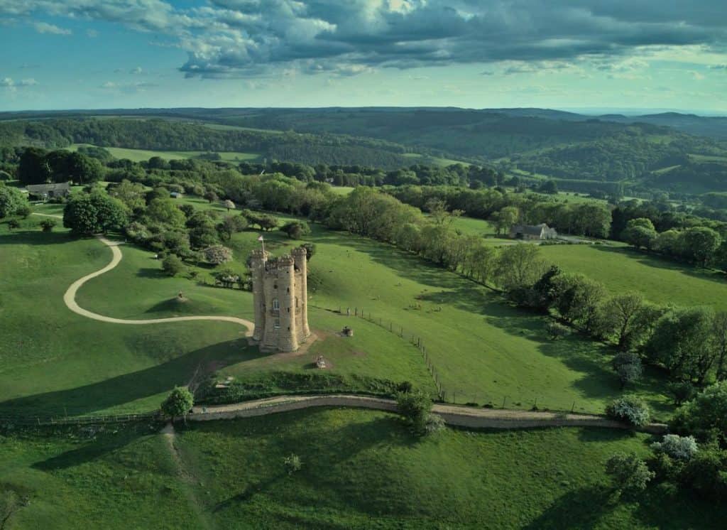 A panoramic view of a stone tower surrounded by vast green meadows and forested hills in the Cotswolds—an enchanting escape for those seeking honeymoon destinations in forest retreats.
