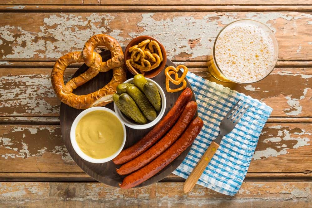 A delicious Bavarian meal featuring pretzels, sausages, mustard, pickles, and a glass of beer, served on a rustic wooden table. Sampling authentic German cuisine like this is one of the best "Things to do in Munich" for any foodie visiting the city.