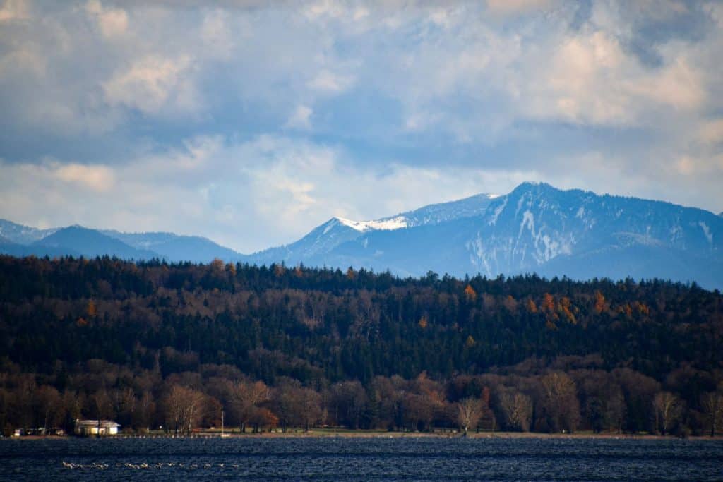 A scenic view of Lake Starnberg in Bavaria, Germany, with a dense forest in autumn colors, calm waters, and the majestic Bavarian Alps in the background. A peaceful getaway just outside the city, visiting this lake is a great addition to "Things to do in Munich" for nature lovers and outdoor enthusiasts.