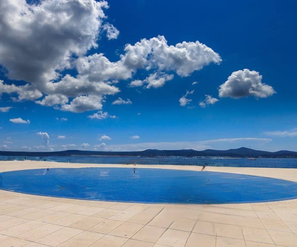 The 'Greeting to the Sun' installation in Zadar, Croatia, reflecting the bright blue sky and scattered clouds, with the Adriatic Sea and distant mountains in the background. This must-see attraction is a perfect stop on any Croatia Summer Itinerary, offering a mesmerizing light show at night powered by solar panels.