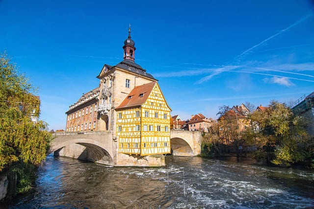 Bamberg, Bavaria, Germany – A stunning view of Bamberg in Bavaria, Germany, featuring the iconic Old Town Hall (Altes Rathaus) standing on a bridge over the Regnitz River. The historic half-timbered building, with its Baroque tower and vibrant yellow-and-white facade, reflects in the flowing water below. This unique landmark is a must-see for Bavaria Germany Travel.