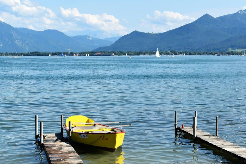 Chiemsee, Bavaria, Germany – A peaceful lakeside view of Chiemsee in Bavaria, Germany, featuring a bright yellow rowboat docked at a wooden pier. The lake stretches toward the distant mountains, with sailboats drifting across the horizon. This picturesque lake is a must-visit for Bavaria Germany Travel.