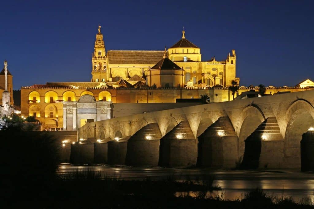  A stunning nighttime view of the illuminated Mosque-Cathedral of Córdoba and the historic Roman Bridge, making it a cultural gem among Cheap Travel Destinations in Europe Summer.