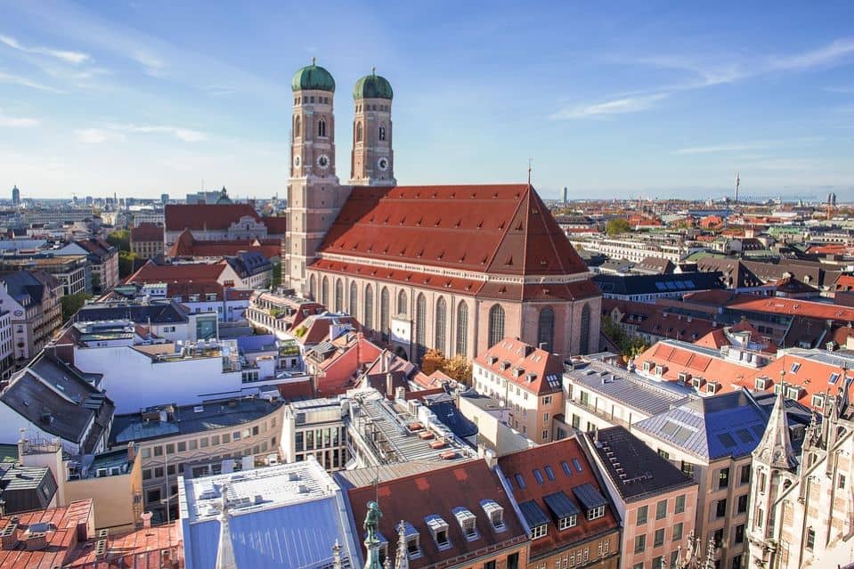 **Munich, Bavaria, Germany** – A stunning aerial view of Munich in **Bavaria, Germany**, featuring the iconic Frauenkirche with its twin green-domed towers rising above the city's historic rooftops. The red-roofed buildings and modern skyline stretch into the distance under a bright blue sky. This vibrant city is a must-visit for Bavaria Germany Travel.