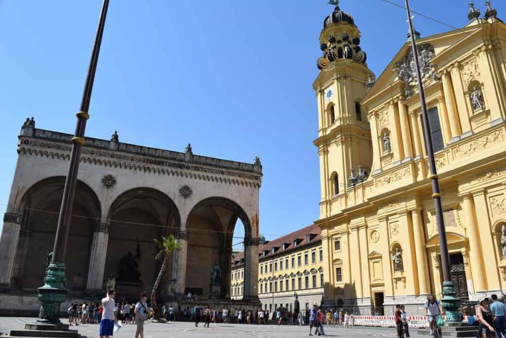 The iconic Odeonsplatz in Munich featuring the grand yellow Theatine Church alongside the Feldherrnhalle, with tourists strolling around under a clear blue sky, a highlight of any Munich 2 Day Itinerary.