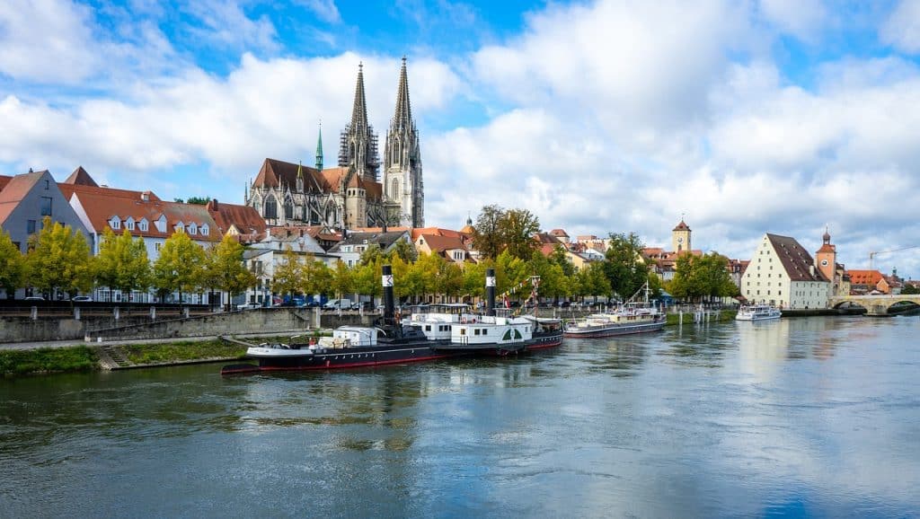 Regensburg, Bavaria, Germany – A stunning view of Regensburg in **Bavaria, Germany**, featuring the majestic Gothic spires of St. Peter’s Cathedral rising above the historic old town. Boats are docked along the Danube River, with a charming riverside promenade lined with colorful buildings. This picturesque city is a must-visit for Bavaria Germany Travel