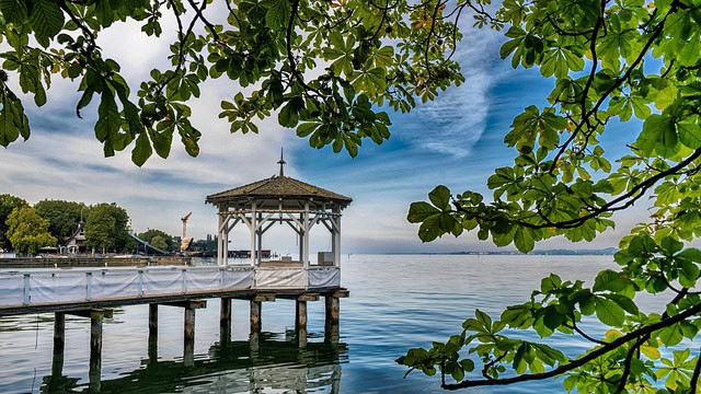 Lake Constance, Bavaria, Germany** – A serene view of Lake Constance in Bavaria, Germany**, featuring a charming wooden gazebo at the end of a pier overlooking the calm water. Lush green leaves frame the scene, with a bright blue sky and wispy clouds reflecting on the lake's surface. This picturesque lakeside retreat is a must-visit for Bavaria Germany Travel