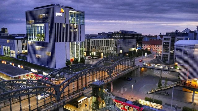 Evening view of the NS-Dokumentationszentrum in Munich, a modern building with sleek, white architecture illuminated by city lights. The surrounding area features contemporary office buildings and a glass-covered pedestrian bridge over a bustling street with trams. As one of the educational highlights among things to do in Munich, this site offers historical insights into the city's past, set against a dramatic, cloudy sky.