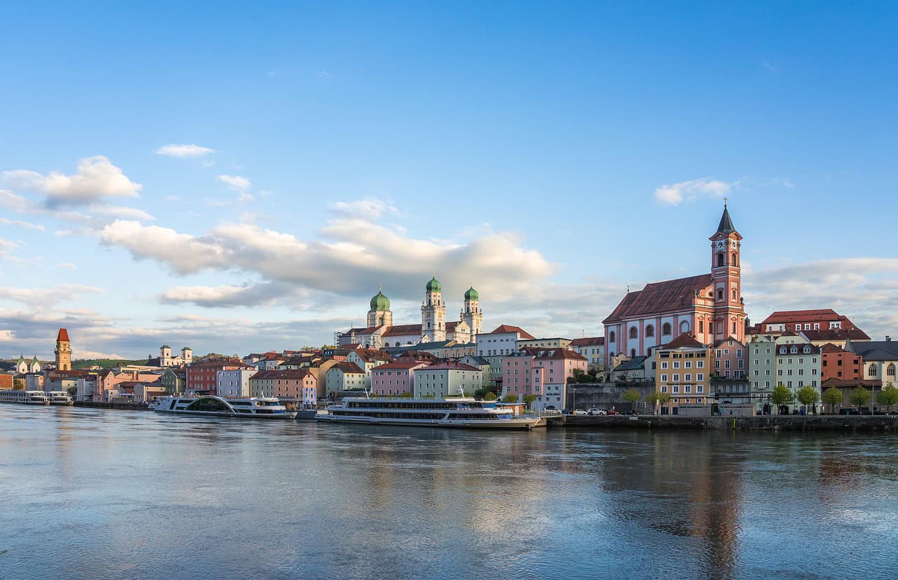 Passau, Bavaria, Germany – A scenic riverside view of Passau in Bavaria, Germany, showcasing colorful historic buildings along the Danube River. The city's famous St. Stephen's Cathedral, with its green-domed towers, stands prominently in the skyline. This picturesque location is a must-visit for Bavaria Germany Travel.