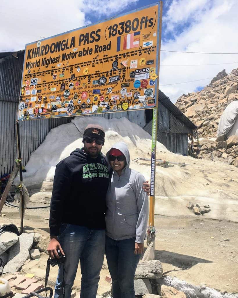 Vrushali and her husband, Akshaya of Couple of Journeys, standing near the  Yellow milestone at Khardung La top, one of the highest motorable roads in the world, surrounded by snow and barren peaks—a high-altitude thrill on a leh ladakh itinerary.