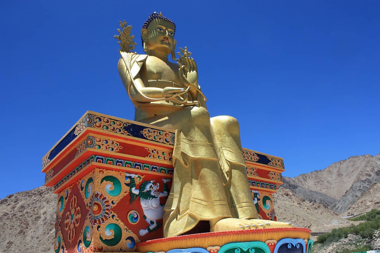 A towering golden Maitreya Buddha statue seated on an ornate, multicolored pedestal with intricate Tibetan patterns, set against a deep blue sky and rocky Nubra Valley mountains—an iconic spiritual site on a leh ladakh itinerary.