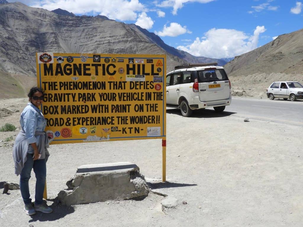Vrushali standing at Magnetic Hill in Ladakh with a signboard inviting visitors to experience the gravity-defying phenomenon, surrounded by stark mountain terrain—a fascinating feature in a leh ladakh itinerary.