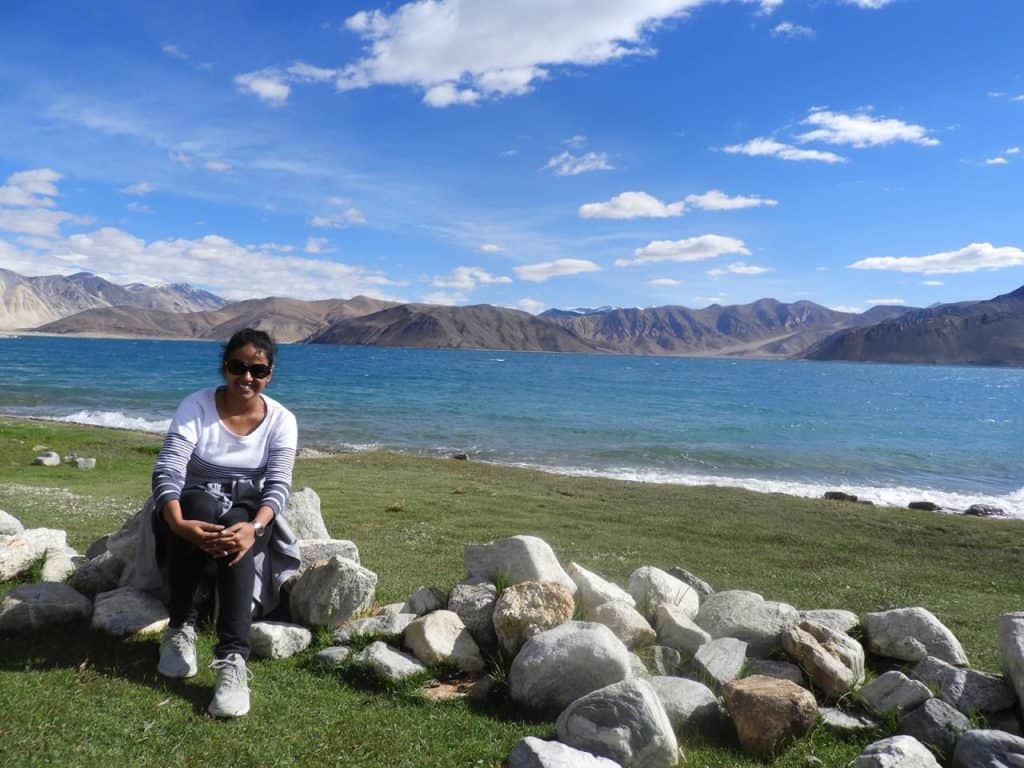 Vrushali sitting at The pristine turquoise waters of Pangong Lake curving along the sandy shoreline, framed by beige mountains and a vibrant blue sky—a top destination in a leh ladakh itinerary.